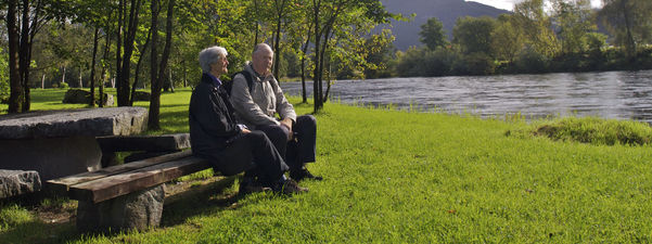Bildet syner ein eldre mann og ei eldre kvinne sit på ein benk ved elva Jølstra i Førde. Det er midt på sommaren, med sol, grønt gras og grønt lauv på trea. Foto: Oskar Andersen