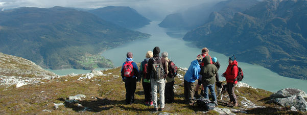 Ni personar kledde for fjelltur står på toppen av fjellet Molden i Luster og ser innover Lustrafjorden. Det er sol, men lågt skydekke. Til venstre ser vi Nes, og til høgre går vegen innover sørsida av Lustrafjorden.