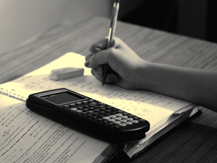 Svart-/kvitt-bilete av ei lærebok i matematikk, ein kalkulator, eit viskelær og ei hand som held hardt i ein blyant. Foto: Steven S., www.flickr.com