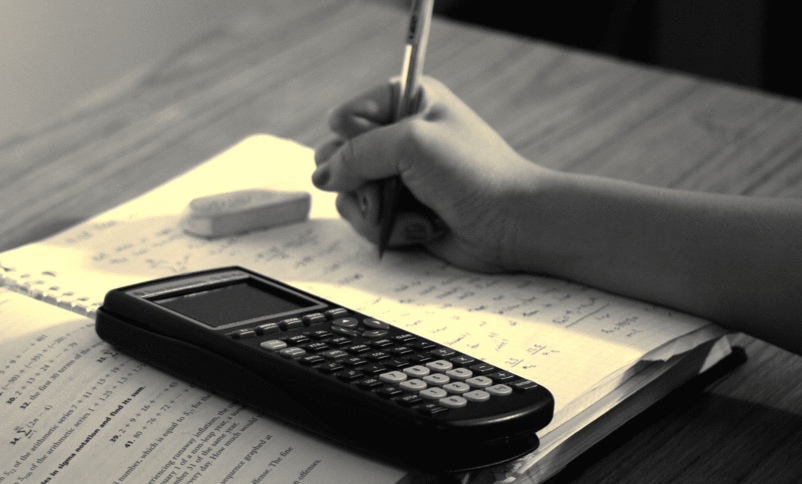 Svart-/kvitt-bilete av ei lærebok i matematikk, ein kalkulator, eit viskelær og ei hand som held hardt i ein blyant. Foto: Steven S., www.flickr.com
