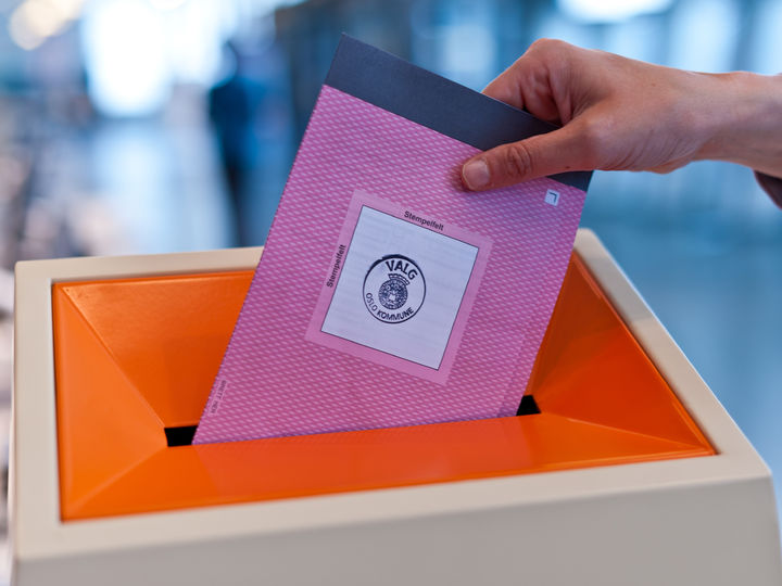Foto: Kommunal- og moderniseringsdepartementet Tore Fjeld Foto som illustrerer val . Biletet syner ei hand som slepper eit rosa valomslag med stemmesetlar ned i ei kvit og oransje valurne. Foto: Kommunal- og moderniseringsdepartementet