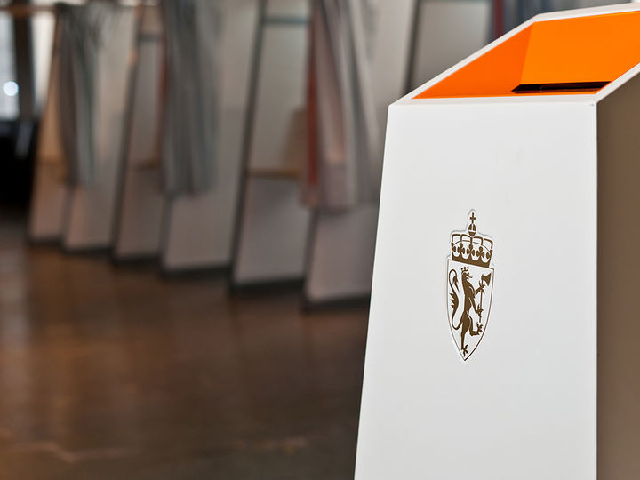 Foto frå eit stemmelokale, der vi ser den kvite og oransje stemmeurna med riksvåpenet i forkant. I bakgrunnen av biletet ser vi valavlukke med stemmesetlar. Foto: Kommunal og moderniseringsdepartementet Tore Fjeld Foto frå eit stemmelokale, der vi ser den kvite og oransje stemmeurna med riksvåpenet i forkant. I bakgrunnen av biletet ser vi valavlukke med stemmesetlar. Foto: Kommunal og moderniseringsdepartementet
