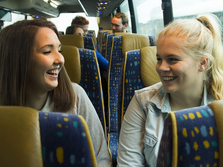 Foto som syner to jenter i ein buss. Dei sit på kvar si side av midtgangen, ei er mørkhåra, og den andre er blond. Foto: A til Å Grafisk Design og Media