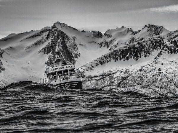 Svart-kvitt bilete av fiskebåt i bølgar med snøkledde fjell i bakgrunnen