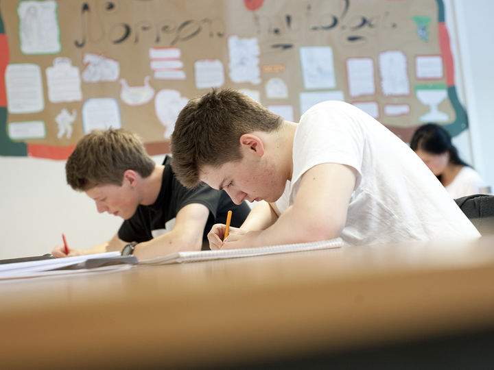 Nærbilete av to gutar som sit bøygde over bøkene i klasserommet og skriv, ein har kvit t-skjorte, den andre svart. I bakgrunnen ser vi elevarbeid på veggen og ei jente. Foto: Hordaland fylkeskommune Nærbilete av to gutar som sit bøygde over bøkene i klasserommet og skriv, ein har kvit t-skjorte, den andre svart. I bakgrunnen ser vi elevarbeid på veggen og ei jente. Foto: Hordaland fylkeskommune
