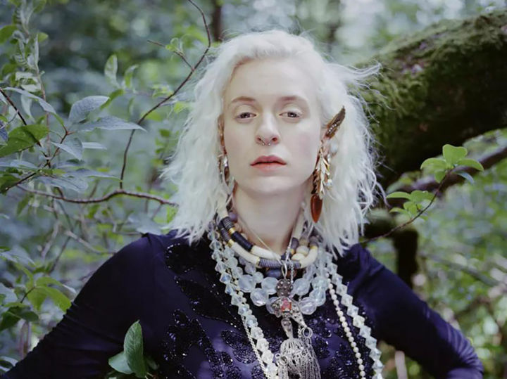 Portrettfoto av artist Thea Hjelmeland. Ho står framfor nokre busker, rett rett i kameraet, har pynt i øyrene, håret og rundt halsen og har på seg ein svart topp. Portrettfoto av artist Thea Hjelmeland. Ho står framfor nokre busker, rett rett i kameraet, har pynt i øyrene, håret og rundt halsen og har på seg ein svart topp.