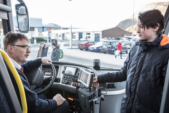 Foto av ein ung gut i samtale med ein bussjåfør på veg inn i bussen. Biletet er teke frå inni bussen og mot frontruta. Utanfor ser vi både bilar og folk på ein parkeringsplass.