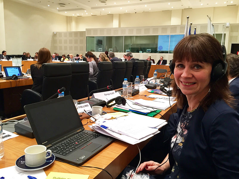 Foto av Jenny Følling på møte i Europarådet sin Congress Monitoring Committe. Biletet er frå møtesalen med mykje folk rundt. Følling ser i kameraet, har øyretelefonar på og pc og papir framføre seg på bordet.