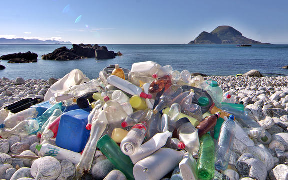 Foto som syner avfall, i hovudsak ulike plastflasker, langs kysten. Vi ser havet og øyer i horisonten, himmelen er blå.