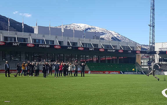Foto frå oppstartssamlinga til det internasjonale prosjektet P-IRIS i april 2017. Biletet er frå Fosshaugane Campus i Sogndal, der deltakarane på samlinga er samla på bana. Ein av dei sparkar ein fotball mot dei andre, sparkar i gang prosjektet.
