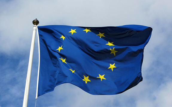Foto av EU-flagget som vaiar mot blå himmel med litt skyer.