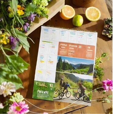 KI generert bilde av en kalender som har et bilde av 3 barn med sykkel. Kalenderen ligger på et bord hvor det også er en appelsin og sitron, samt sommerblomster.