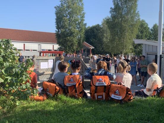 1. klasse elever med oransje skolesekk sitter på gressplenen.