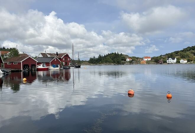 Bilde tatt på sjøen. En ser to oransje dobber, røde båthus til venstre  og hvite hus til høyre i bilde