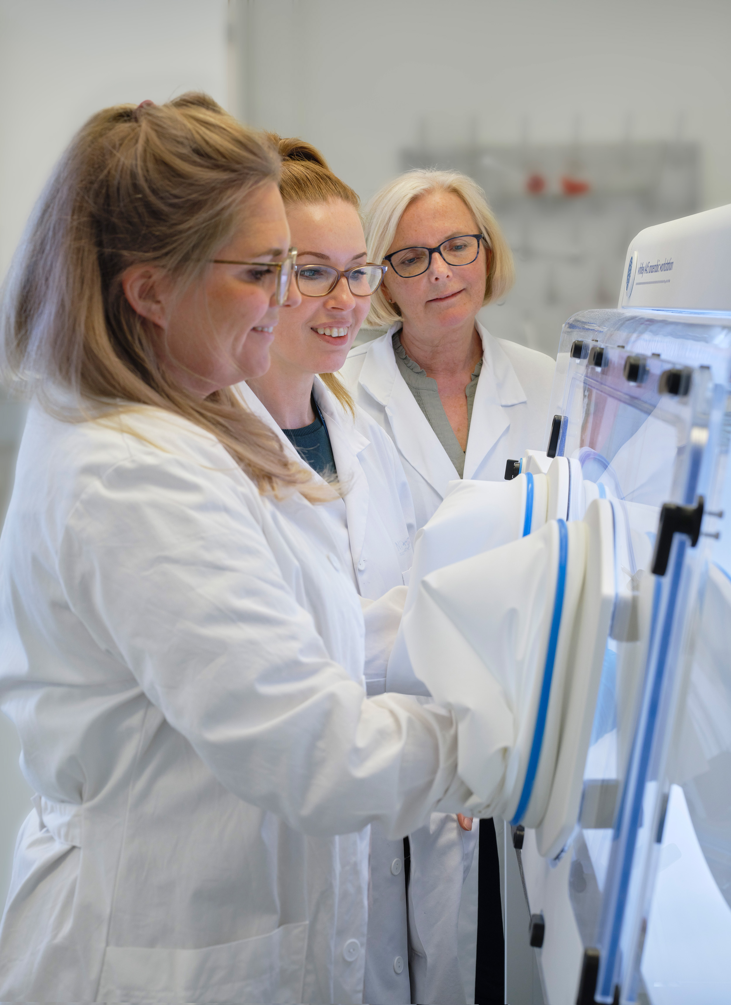 Picture of visiting scientist Helene Rygvold and NIOM employees Maria Pain and Inger Sofie Dragland standing by the anarobe bench in the NIOM lab